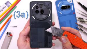 Thumbnail of a smartphone with a cracked screen, surrounded by various tools used for durability testing, including a utility knife and a screwdriver, on a wooden surface.